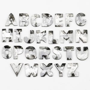 Pode incluir: Um conjunto completo de alfabeto de letras prateadas e facetadas. Cada letra tem uma forma e tamanho únicos, com uma superfície texturizada e reflexiva. As letras estão dispostas sobre um fundo branco.