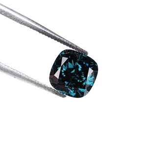 May include: A cushion-cut, teal-colored gemstone is held by a pair of silver tweezers against a white background. The gemstone has multiple facets that reflect light, creating a sparkling effect. The overall impression is one of luxury and elegance.