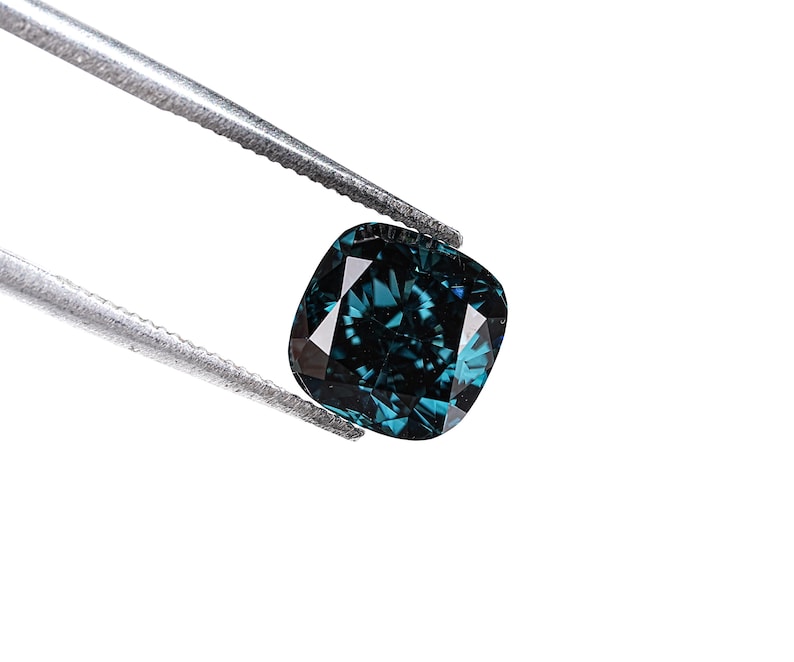 May include: A cushion-cut, teal-colored gemstone is held by a pair of silver tweezers against a white background. The gemstone has multiple facets that reflect light, creating a sparkling effect. The overall impression is one of luxury and elegance.