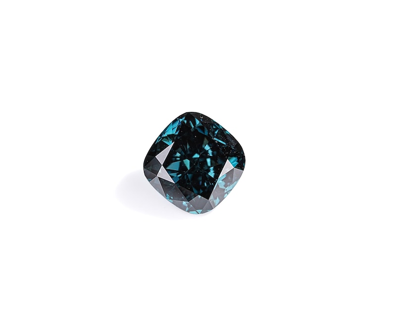 May include: A cushion-cut, teal-colored gemstone with a faceted surface. The gem is set against a white background, highlighting its deep color and intricate cuts. The gemstone's shape is square with rounded corners.
