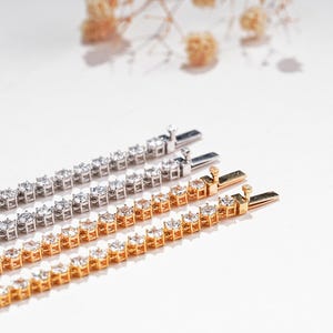 K&ouml;nnte beinhalten: Nahaufnahme von drei Tennisarmb&auml;ndern. Zwei sind goldfarben und eines silberfarben. Jedes Armband hat eine Reihe klarer, funkelnder Edelsteine. Die Armb&auml;nder werden auf einer wei&szlig;en Oberfl&auml;che mit weicher Beleuchtung pr&auml;sentiert.