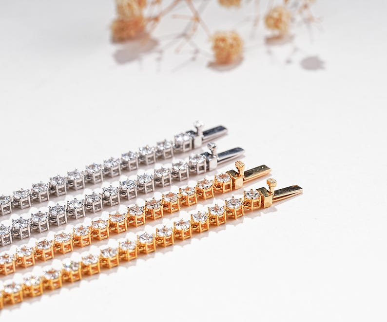 K&ouml;nnte beinhalten: Nahaufnahme von drei Tennisarmb&auml;ndern. Zwei sind goldfarben und eines silberfarben. Jedes Armband hat eine Reihe klarer, funkelnder Edelsteine. Die Armb&auml;nder werden auf einer wei&szlig;en Oberfl&auml;che mit weicher Beleuchtung pr&auml;sentiert.