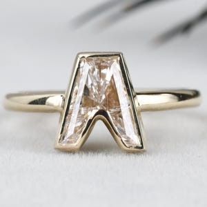 Könnte beinhalten: Ein goldener Ring mit einem hellfarbenen Edelstein in Form des Buchstabens "A". Der Stein ist in einer Lünettenfassung gefasst. Der Ring hat ein schmales Band. Der Hintergrund ist weich und neutral. Ein Schmuckstück.