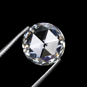 May include: A round, clear gemstone with multiple facets, held by silver tweezers against a black background. The gemstone reflects light, creating a sparkling effect. The image highlights the clarity and cut of the diamond.