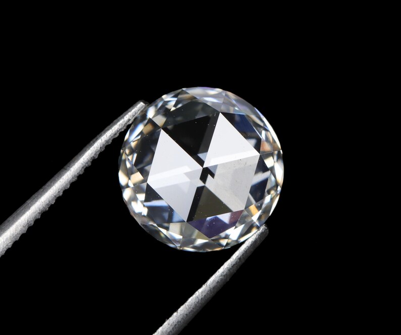May include: A round, clear gemstone with multiple facets, held by silver tweezers against a black background. The gemstone reflects light, creating a sparkling effect. The image highlights the clarity and cut of the diamond.