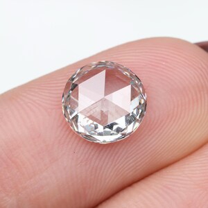 May include: A clear, round-cut gemstone with a faceted surface. The gem is held between two fingers, showcasing its clarity and geometric design. The gemstone appears to be a diamond or similar precious stone.