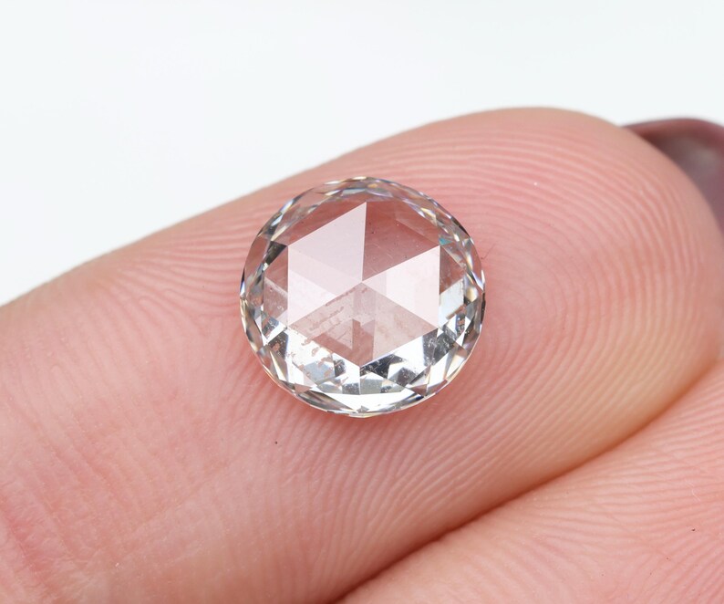 May include: A clear, round-cut gemstone with a faceted surface. The gem is held between two fingers, showcasing its clarity and geometric design. The gemstone appears to be a diamond or similar precious stone.