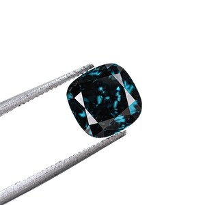 May include: A cushion-cut, teal-colored gemstone held by silver tweezers against a white background. The gemstone has a faceted surface that reflects light, creating a sparkling effect. The overall shape is square with rounded corners.