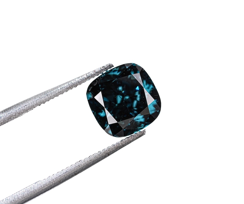 May include: A cushion-cut, teal-colored gemstone held by silver tweezers against a white background. The gemstone has a faceted surface that reflects light, creating a sparkling effect. The overall shape is square with rounded corners.