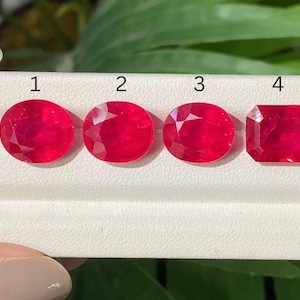 May include: Four vibrant red gemstones arranged in a row on a white display stand. The first three stones are oval-shaped, while the fourth stone is rectangular with cut corners. Each stone is labeled with a black number from one to four above it.