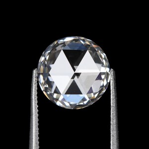 May include: A clear, round-cut diamond held by silver tweezers against a black background. The diamond's facets reflect light, creating a sparkling effect. The gem is a brilliant, colorless stone, showcasing its clarity and cut.