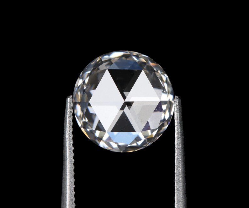 May include: A clear, round-cut diamond held by silver tweezers against a black background. The diamond's facets reflect light, creating a sparkling effect. The gem is a brilliant, colorless stone, showcasing its clarity and cut.