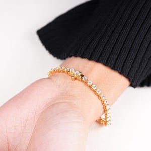 K&ouml;nnte beinhalten: Ein goldfarbenes Armband mit klaren, runden Edelsteinen wird an einem Handgelenk gezeigt. Das Armband ist in Goldmetall gefasst und weist eine Reihe von gleichm&auml;&szlig;ig verteilten, funkelnden Steinen auf. Das Handgelenk liegt vor einem schwarzen Pullover, mit wei&szlig;em Hintergrund.