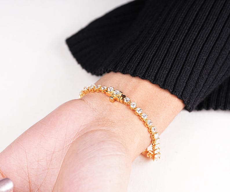 K&ouml;nnte beinhalten: Ein goldfarbenes Armband mit klaren, runden Edelsteinen wird an einem Handgelenk gezeigt. Das Armband ist in Goldmetall gefasst und weist eine Reihe von gleichm&auml;&szlig;ig verteilten, funkelnden Steinen auf. Das Handgelenk liegt vor einem schwarzen Pullover, mit wei&szlig;em Hintergrund.