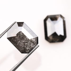 Könnte beinhalten: Zwei roh geschliffene, smaragdformige Diamanten. Einer wird von einer Pinzette gehalten, die seine Facetten und das dunkle, gesprenkelte Innere zeigt. Der andere Diamant ist im Hintergrund unscharf. Die Diamanten sind vor einem weißen Hintergrund platziert.