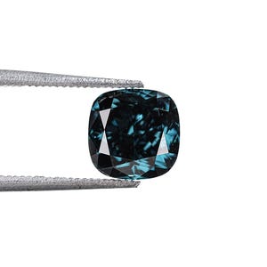 May include: A cushion-cut, teal-colored gemstone held by tweezers against a white background. The gem has a square shape with rounded corners and facets that reflect light. The color is a deep teal with hints of blue and green.