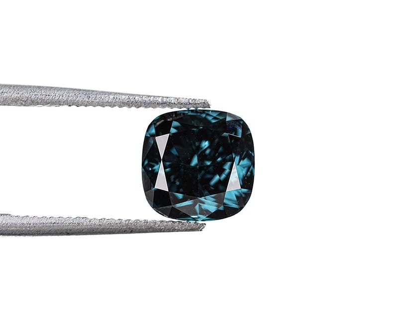 May include: A cushion-cut, teal-colored gemstone held by tweezers against a white background. The gem has a square shape with rounded corners and facets that reflect light. The color is a deep teal with hints of blue and green.