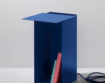 Op de afbeelding: Een blauwe metalen bijzettafel met een plank en een scharnierend blad. De plank bevat drie boeken, een roze, een blauwe en een met een zwarte kaft.