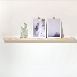 Op de afbeelding: Drie heldere glazen flessen staan op een licht houten plank met twee zwart-witte ingelijste foto's van mistige landschappen erboven.