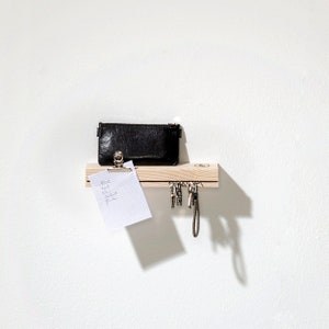 Könnte beinhalten: Ein Schlüsselbrett aus Holz mit einem schwarzen Lederportemonnaie, einem weißen Notizblock mit einem Clip und einem Schlüsselbund, der an Haken hängt. Das Brett ist an einer weißen Wand montiert.