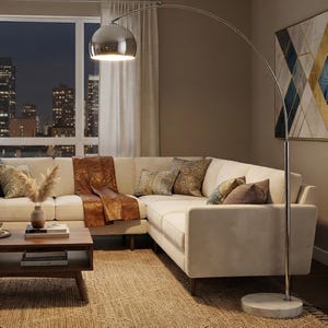 May include: A tall, arched floor lamp with a polished chrome finish, a large dome-shaped shade, and a circular white marble base. The lamp is positioned next to a cream-colored sectional sofa, casting a warm, bright light downward from the shade.