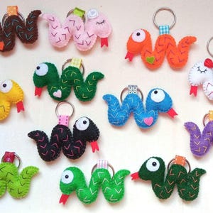 Könnte beinhalten: Eine Sammlung von 10 Schlüsselanhängern aus Filz in verschiedenen Farben, darunter Grün, Blau, Lila, Orange, Pink, Gelb und Braun. Jede Schlange hat ein süßes Gesicht und ist mit einem Zickzackmuster genäht.