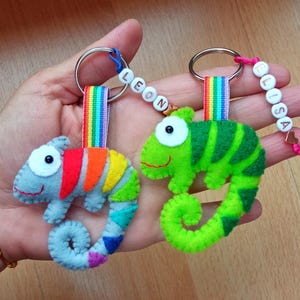 Peut inclure: Deux porte-clés faits à la main en forme de caméléon. L'un est gris avec une queue arc-en-ciel, l'autre est vert avec un motif rayé. Chacun a une sangle arc-en-ciel et un anneau métallique. Un porte-clés porte le nom "LEON" et l'autre le nom "ELISA".