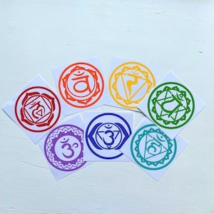 May include: Seven colourful round stickers with different chakra symbols and the Sanskrit word "Om" in the centre. The stickers are arranged in a circle on a white wooden surface.