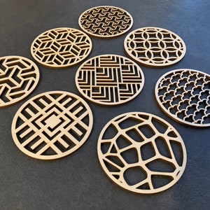 Puede incluir: Ocho posavasos redondos de madera con diferentes diseños geométricos. Los posavasos están hechos de madera marrón claro y tienen un patrón cortado con láser.