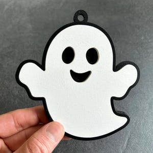 May include: A wooden Halloween ghost decoration. The ghost is white with a black outline and features a smiling face with black eyes and mouth. It has a small hole at the top for hanging.