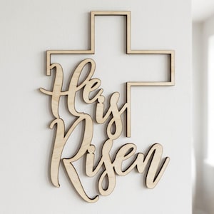 May include: A wooden cross with the words "He is Risen" cut out in a cursive font. The cross is a simple, geometric shape, and the text is layered over the bottom half of the cross. The wood has a light, natural finish.
