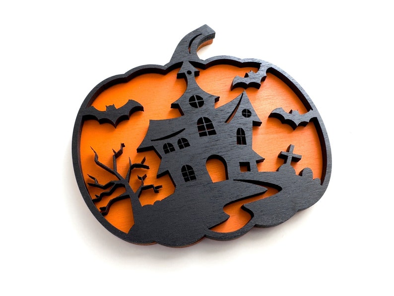 May include: A black wooden Halloween decoration in the shape of a pumpkin. The pumpkin features a haunted house, bats, a tree, and a graveyard silhouette against an orange background. A festive, seasonal item.
