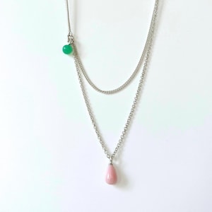 Op de afbeelding: Een gelaagde zilveren ketting met een kleine groene kraal en een roze, druppelvormige hanger. De ketting wordt gepresenteerd tegen een witte achtergrond, die het delicate ontwerp en het kleurcontrast benadrukt.