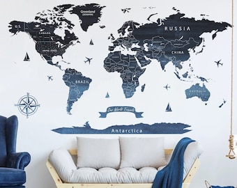 May include: A living room interior with a large world map wall decal in blue and white. The map features continents and countries with names in white text. The room also includes a blue armchair, a light wood sofa with white cushions, a blue throw blanket, a wicker basket, and a small wooden shelf with a sign that reads "Good Morning".