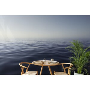 May include: A wooden table with two chairs and a plant sits in front of a blue ocean with a hazy sky.