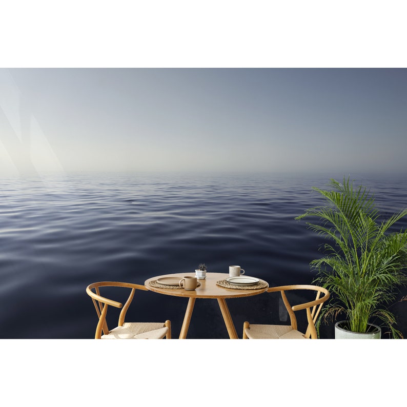 May include: A wooden table with two chairs and a plant sits in front of a blue ocean with a hazy sky.