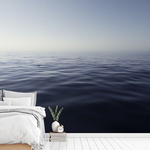 May include: A white bed with white bedding and two pillows sits in front of a wall with a photo of a calm, blue ocean. A small green plant sits on a stack of books next to a white alarm clock.