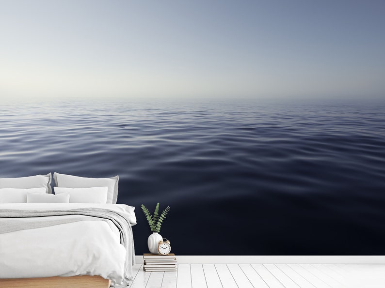 May include: A white bed with white bedding and two pillows sits in front of a wall with a photo of a calm, blue ocean. A small green plant sits on a stack of books next to a white alarm clock.