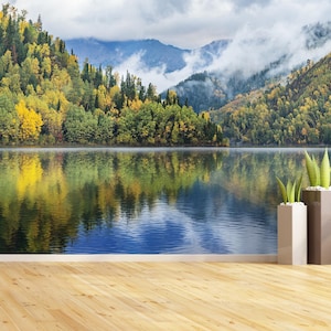 Può includere: Un paesaggio sereno con un lago che riflette gli alberi autunnali e le montagne. Gli alberi mostrano colori gialli e verdi, con un cielo nuvoloso. Due fioriere decorative con piante verdi in primo piano.