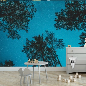 May include: A whimsical nursery scene featuring a starry night sky wallpaper with silhouetted trees. A gray dresser, small table, and chair are in the foreground. A bunny-shaped lamp and wooden toy train add to the playful atmosphere. A banner reads "Dream Big".