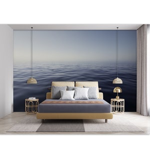 May include: A bedroom interior with a bed, two end tables, and two hanging lamps. The wall behind the bed features a mural of a calm, blue ocean with a hazy horizon.