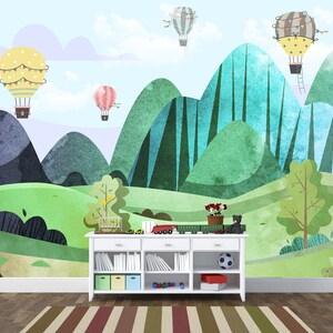 K&ouml;nnte beinhalten: Ein wei&szlig;es B&uuml;cherregal mit einem gr&uuml;nen, blauen und gelben Berglandschaft-Wandbild mit Hei&szlig;luftballons. Das B&uuml;cherregal hat vier Regalbretter und ist mit einem Fu&szlig;ball, einem Spielzeugzug und einer Topfpflanze dekoriert.