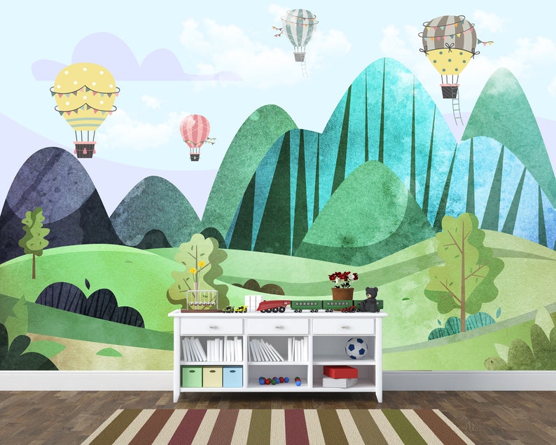 K&ouml;nnte beinhalten: Ein wei&szlig;es B&uuml;cherregal mit einem gr&uuml;nen, blauen und gelben Berglandschaft-Wandbild mit Hei&szlig;luftballons. Das B&uuml;cherregal hat vier Regalbretter und ist mit einem Fu&szlig;ball, einem Spielzeugzug und einer Topfpflanze dekoriert.