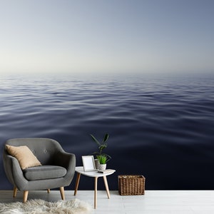 May include: A gray armchair with a tan pillow sits in front of a wall with a photo of a calm, blue ocean. A small white table with a potted plant and a picture frame sits next to the chair. A wicker basket sits on the floor.
