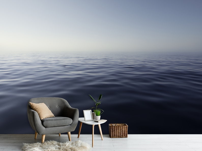May include: A gray armchair with a tan pillow sits in front of a wall with a photo of a calm, blue ocean. A small white table with a potted plant and a picture frame sits next to the chair. A wicker basket sits on the floor.