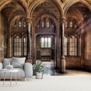 May include: An interior shot of a stone building with arched ceilings and large windows. A gray armchair, a white side table with a vase, and a potted plant are in the foreground. The building has a historic, gothic architectural style.