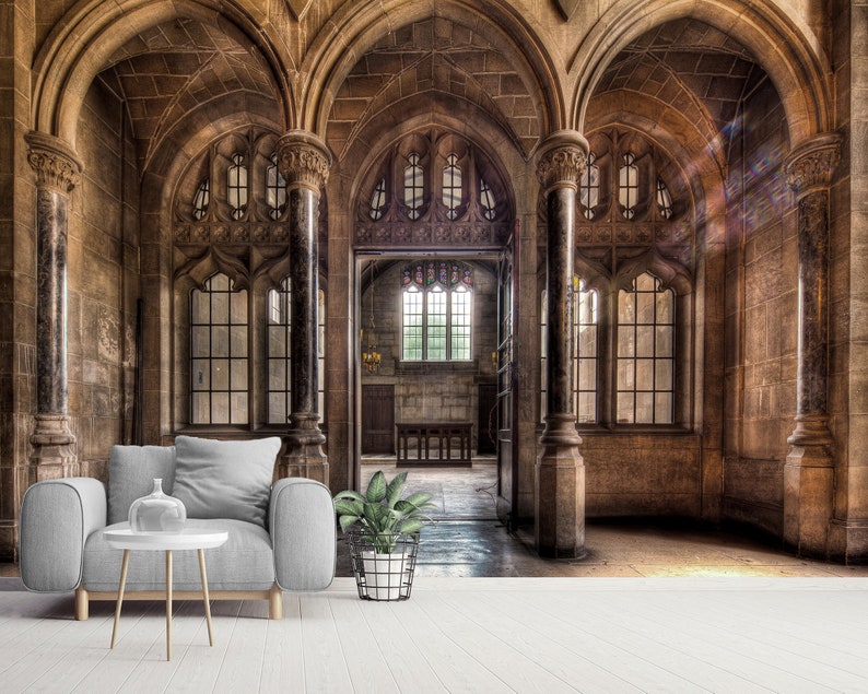 May include: An interior shot of a stone building with arched ceilings and large windows. A gray armchair, a white side table with a vase, and a potted plant are in the foreground. The building has a historic, gothic architectural style.