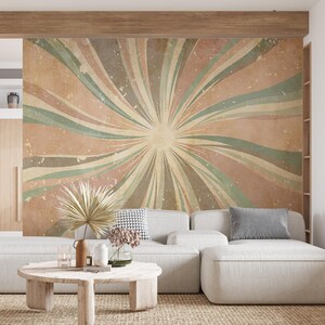 Op de afbeelding: Een woonkamer met een groot abstract muurschildering met een zonnestraalontwerp in gedempte tinten perzik, groen en beige. Een lichtgekleurde hoekbank, een ronde houten salontafel en decoratieve items maken de ruimte compleet.