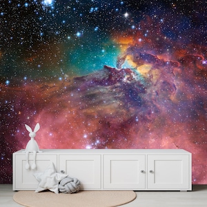 Puede incluir: Una figurita de conejo blanco se sienta en una c&oacute;moda blanca frente a un mural de pared que representa una nebulosa colorida con estrellas.