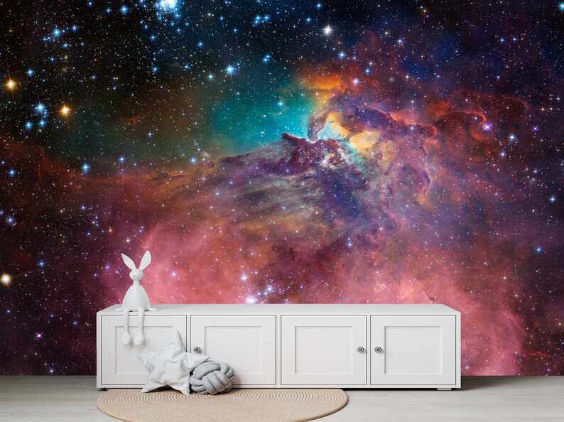 Puede incluir: Una figurita de conejo blanco se sienta en una c&oacute;moda blanca frente a un mural de pared que representa una nebulosa colorida con estrellas.